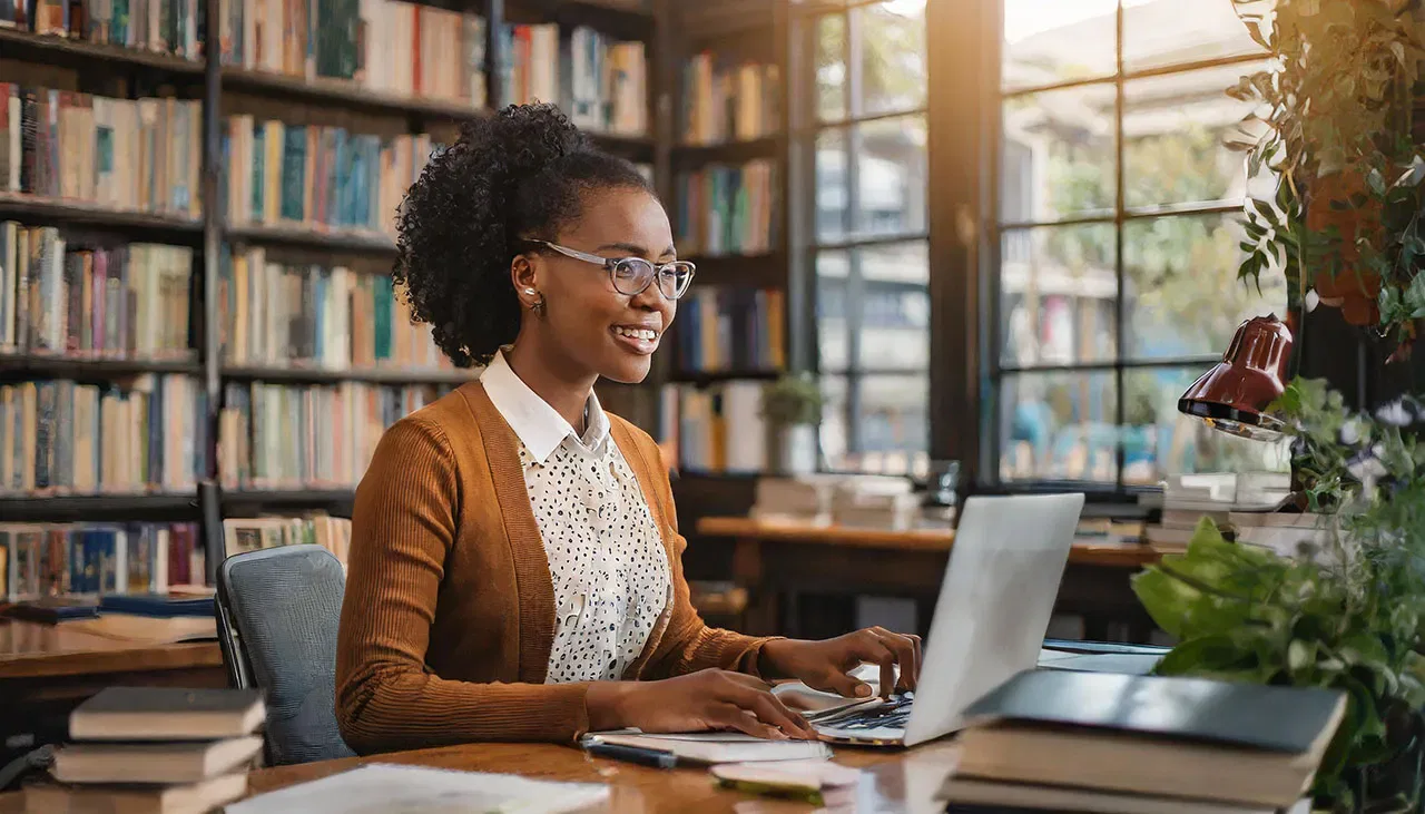 A cheerful young author with glasses using a laptop at a desk in a library filled with books as she works on elements of her author platform.