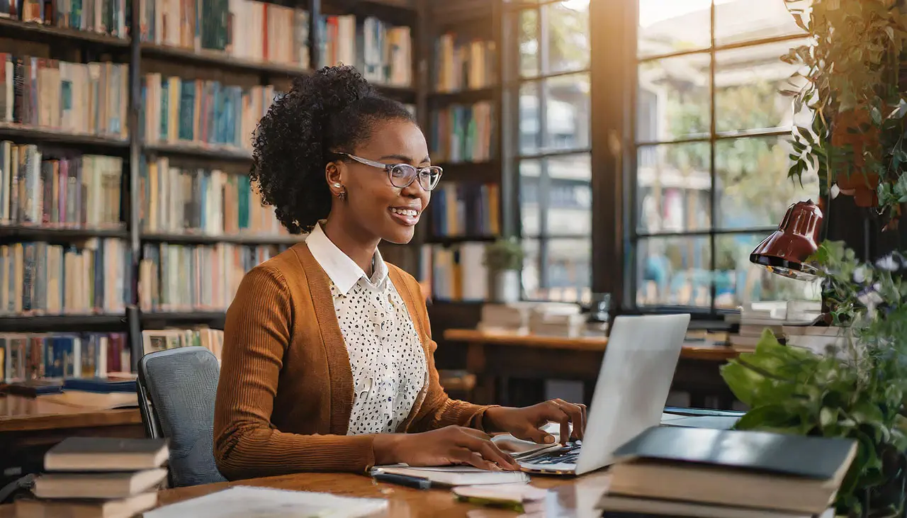 A cheerful young author with glasses using a laptop at a desk in a library filled with books as she works on elements of her author platform.